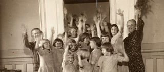 En grupp kvinnor och barn i rutiga klänningar står i en dörröppning och vinkar