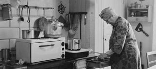 En elektrisk spis med stor kaffekittel på i ett äldre, omodernt kök. En kvinna skär upp någonting