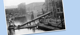 Ett stort rör är på väg att sänkas ned i vattnet framför stora hus.