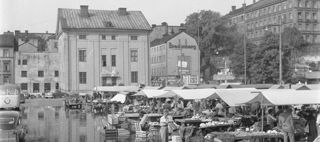 I förgrunden marknadsstånd och parkerade bilar, i bakgrunden byggnader och grönska