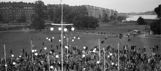 Upplyst dansbana med många människor i en  park med träd och byggnader i bakgrunden