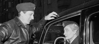 En stående manlig taxichaufför lutad mot en bil. En sittande kvinnlig taxichaufför i bilen.