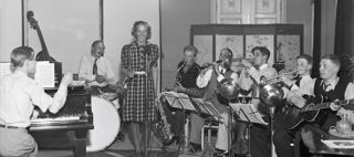 Sångerskan Alice Babs sjunger i en mikrofon och runt omkring henne sitter musiker som spelar olika instrument.