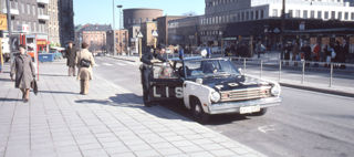 En polis står i dörren till en polisbil vid en trottoar där människor går förbi.