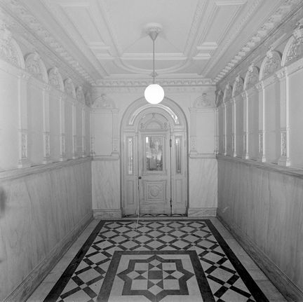 Entréhallen till ett större flerfamiljshus. I änden en port med rundad överkant. golvet är mönsterlagt med rutmönster och har en stjärna i mitten. I taket hänger en pendellampa med en enkel, lysande glob.