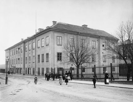 Svartvit bild med en stor byggnad i centrum. Utanför på gatan syns barn vid ett staket, samt två träd utan löv. Barnen är klädda i hatt eller mössa på huvudet.