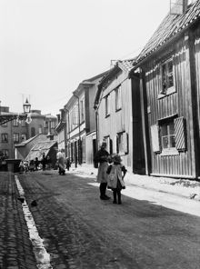 Folkliv på Breda Gatan 2-6. Breda Gatan går mot sydväst till Östra Varvsgatan i fonden. Dåvarande kv. Kyrkogården (t.h.), nu kv. Krigsmanskassan. Kv. Bergsklippan i fonden