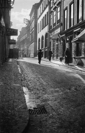 En smal stadsgata kantas av stenhus i två till fyra våningar. I snömodden går tre kvinnor klädda i långa kjolar, kappa och bredbrättad hatt. Solen skiner.