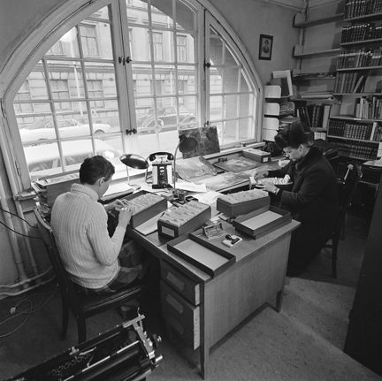 Kontorsinteriör. Vid ett skrivbord mot ett stort fönster sitter en man och en kvinna och arbetar. Manne har en pälsmössa på huvudet. Båda letar efter något i kartotekslådor. 