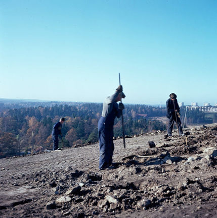 Tre män står på en kulle och arbetar med spett. Milsvid utsikt.