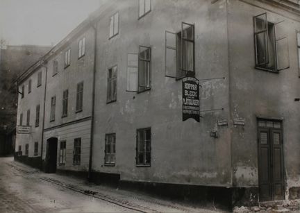 Ett äldre stenhus i två-tre våningar ligger i en sluttande backe. Från fasaden hänger en butiksskylt som berättar att det finns ett plåtslageri i byggnaden.