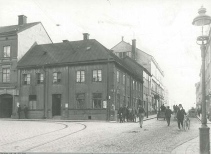 Folkliv framför trähuset där krogen låg. Här skymtar en polis, en man med cykel, ett par kärror, med mera.