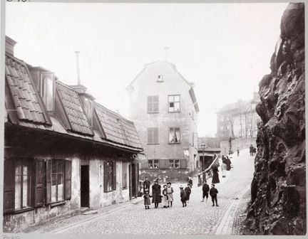 En kullerstensbelagd gata går uppför ett berg. Gatan kantas på ena sidan av låga trähus och ett större, högre, stenhus. På andra sidan en kal och brant bergvägg. Mitt i gatan står ett tiotal barn.