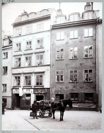 Två äldre stenhus med ett torg belagt med gatsten framför. På torget står en häst med vagn. Intill vagnen står en man med en väska i handen.