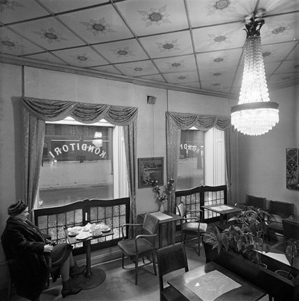 Interiör från ett äldre kafé. Taket är av dekorerade glasplattor. I fönstret står skärmar av glas och fungerar både som dekor och insynsskydd. I det ena hörnet sitter en kvinna med hatt och kappa på, i det andra en man som äter på något. Små bord och stolar och en blomlåda i mitten. En 30- eller 40-talsspegel på väggen.