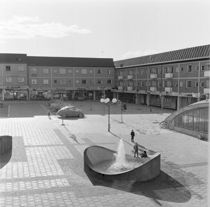 Torg med fontän och parkerade bilar.