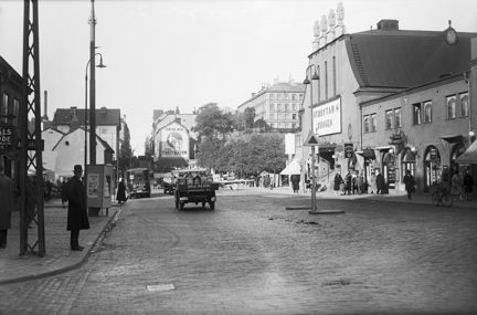 En bred stadsgata kantas av stenhus i ett par våningar. Till höger i bild ligger en stor biografbyggnad med slät fasad och ett entréparti som kröns av fyra stora urnor. I bottenvåningen ligger flera butiker. Människor promenerar förbi och på gatan kör en buss och en mindre lastbil med mjölkkannor.
