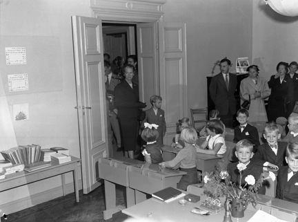 Klassrum med barn i skolbänkar och stående vuxna.