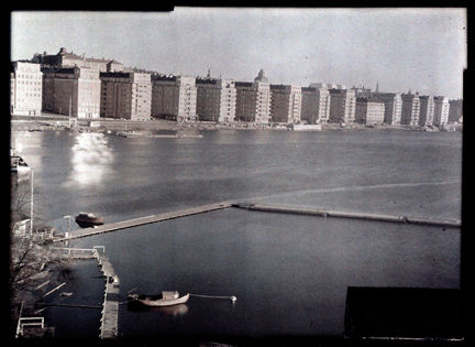 Längs ett större vatten syns en rad med flerfamiljshus i funktionalistisk stil. Från land, ungefär där fotografen som tagit bilden står, går det ut flera långa båtbryggor.