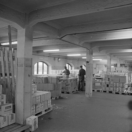 En stor lokal med pelare och pallar med kartongen. I bakgrunden syns två män vid ett fönster.