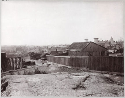 Svart-vit bild med utsikt av bebyggelse. I förgrunden klippor och i centrum ett lågt plank. Bakom planket syns några enklare trähus. I fonden till höger Katarina kyrka.