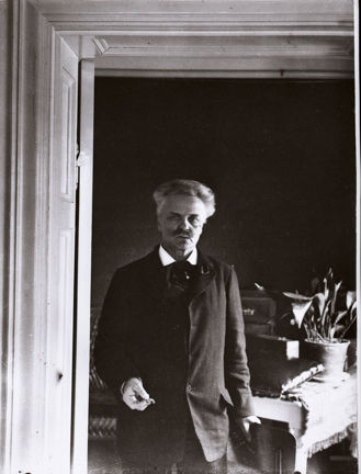 August Strindberg står i en dörröppning, iklädd mörk kostym och kravatt och med en cigarett i handen. I bakgrunden ett bord med duk, en lerkruka med en växt.