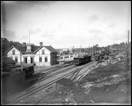 Intill ett spårområde ligger en ljusmålad äldre järnvägsstationsbyggnad av trä. Till vänster om den anas en husrad med höga stenhus från slutet av 1800-talet. 