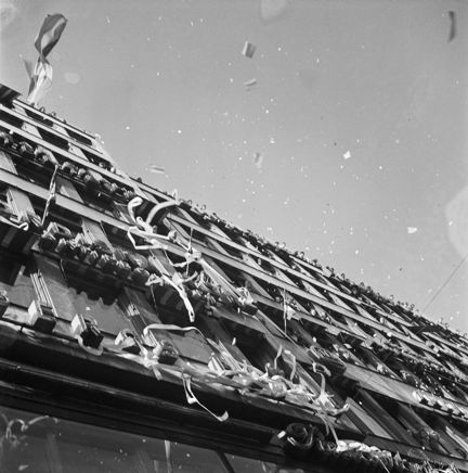 Fredsdagen den 7:e maj 1945 i Stockholm.
Vy upp längs husfasad till Kungsgatan 18 med människor i fönstren som kastar ut papper och girlanger för att fira freden.
