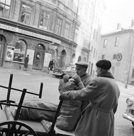 i förgrunden syns en kärra med två vedsäckar. En man i keps håller i en av säckarna. Bredvid står en man i basker och rock med pälskrage. I bakgrunden syns Sundbergs konditori och Norra Bankohuset.