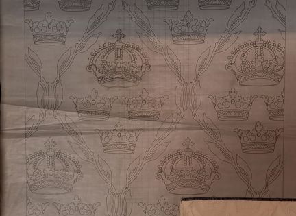 Ett tunt mönsterpapper med ett mönster ritat på. Mönstret föreställer tre små kronor och en större, sluten krona ovanför de tre små. Runt de fyra kronorna är det ritat en ram av löv. Pappret är rutat på några ställen.