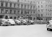 Parkerade bilar på Brunkebergstorg vid Brunkebergspumpen. Herkulesgatan mynnar vid cyklarna. Trådbussledningar hänger i luften. Kv. Åskslaget är nuvarande kv. Trollhättan med Gallerian