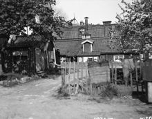Äppelträdet blommar på gårdssidan vid Yttersta Tvärgränd 4. Taket bakom skorstenen hör till huset vid Yttersta Tvärgränd 3. I fonden går några personer på Skinnarviksberget