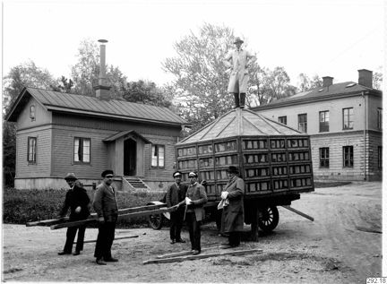 Svartvitt fotografi av fem män med en kärra med den inpackade tempelmodellen. Ovanpå står en man. I bakgrunden hus och träd.