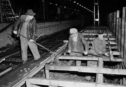 Tre män arbetar med att ta bort räls i ett tunnelbanespår. Två av männen arbetar med spett, en man arbetar med slägga. Två av männen bär keps, den tredje hatt och läderjacka. I förgrunden syns de träställningar som rälsen vilar på.
