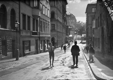 Det ser ut att vara vår och solen lyser starkt ned på en stadsgata. Mitt i gatan står en pojke i keps. I närheten går ett par män. Gatan kantas av större stenhus i ett par våningar och i fonden anas en grönskande kulle..