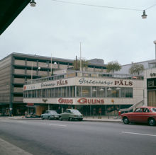 Vy snett över Mäster Samuelsgatan mot affärerna Gulins och Stridsbergs Päls i kv. Beridarebanan. Terrass med träd på byggnadens tak