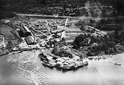 Flygbild över en hamn med båtar, upplag av timmer. I bakgrunden ett skogsbryn, väg och spårvagnslinje.