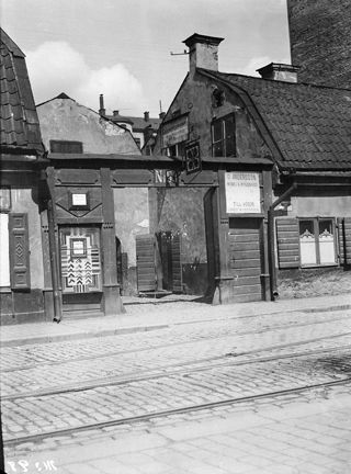 Gata med spårvagnsspår. Tvåvåningshus med fönsterluckor i trä. En vid öppning leder mot gården.