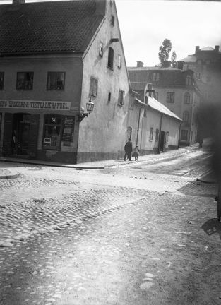 I en gatukorsning ligger ett stenhus av äldre karaktär och i två våningar. Gatan är belagd med gatsten och grus. Två barn går förbi huset. På fasaden sitter flera butiksskyltar.