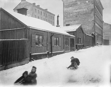 Några barn åker kälke nerför en backe. Bakom ligger gammal trähusbebyggelse samt några flerfamiljshus .