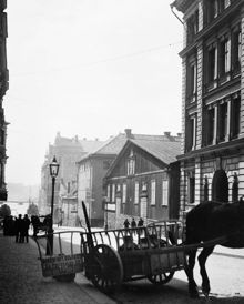 En hästkärra med mjölkkrukor står vid Grevgatan 5 (nu 15). Skylten på kärran hänvisar till S:t Paulsgatan 22. Efter trähuset t.h. ligger hörnet av Riddargatan. I fonden söderut skymtar Nybroviken
