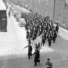 Svea Livgarde på Riddargatan i hörnet av Artillerigatan på väg till Riksdagens högtidliga öppnande. Uniformerna är av 1820-tals modell. På fanan markeras fältslagen