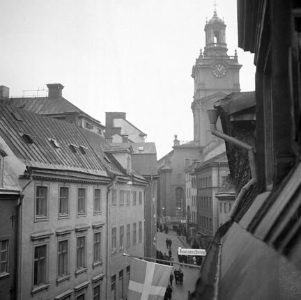 En flagga sticker ut från fasaden. Folkliv nere på gatan.