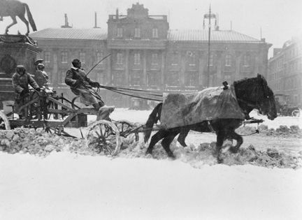 Två hästar drar en vagn med snöplog. På vagnen finns tre personer. I bakgrunden syns Arvsfurstens palats som är Sveriges Utrikesdepartement sedan 1906.