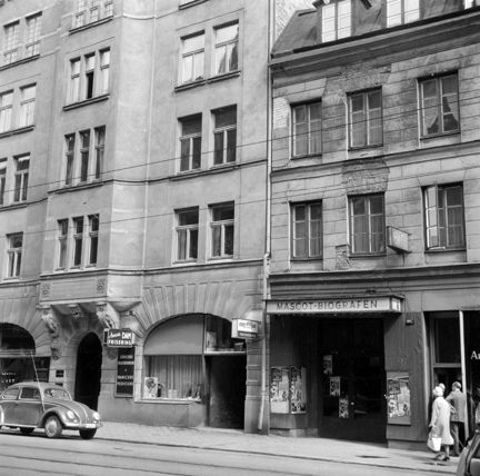 Gatumiljö. Biografentré, skyltfönster, parkerad bil, förbipasserande.