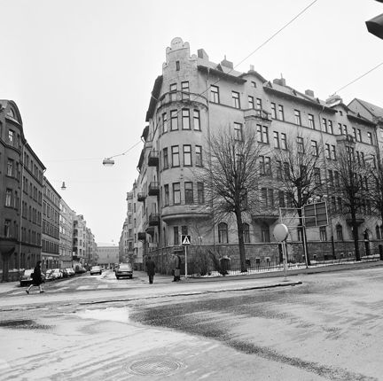 En gatukorsning med flerbostadshus. någar bilar står parkerade, några perosner går över gatan.