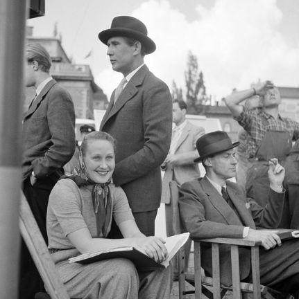 personer vid filminspelning sitter på stolar, skådespelaren Georg Rydeberg står bakom i kostym och hatt.