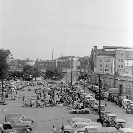 På en stor öppen plats står flera bilar parkerade. Platsen är fylld av trälådor med frukt och grönsaker. Torghandel pågår. I bakgrunden syns flera stora stenhus och stadsmiljö. 