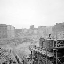 Beridarbansgatan 16 vid hörnet av Hamngatan 38 under rivning (kv. Hoven). Beridarbansgatan 18, kv. Stigbygeln är rivet. Mäster Samuelsgatan går på en bro. Bakom Konstfackskolan skymtar Konserthuset