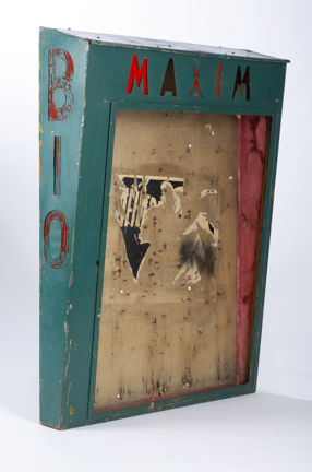 Ett skåp fotat snett från vänster med mörkgrön ram och texten "Maxim" i överkant. Till vänster om ramen står texten: "Bio."